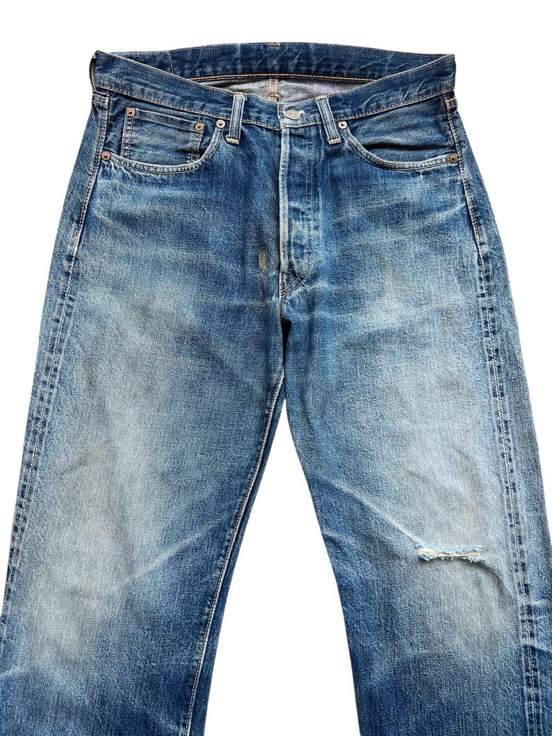 以下が含まれることがあります： ボタンフライとクラシックな5ポケットデザインの、色あせたブルーデニムジーンズ。右脚に小さな破れなど、使用感があり、ヴィンテージ感のあるジーンズです。