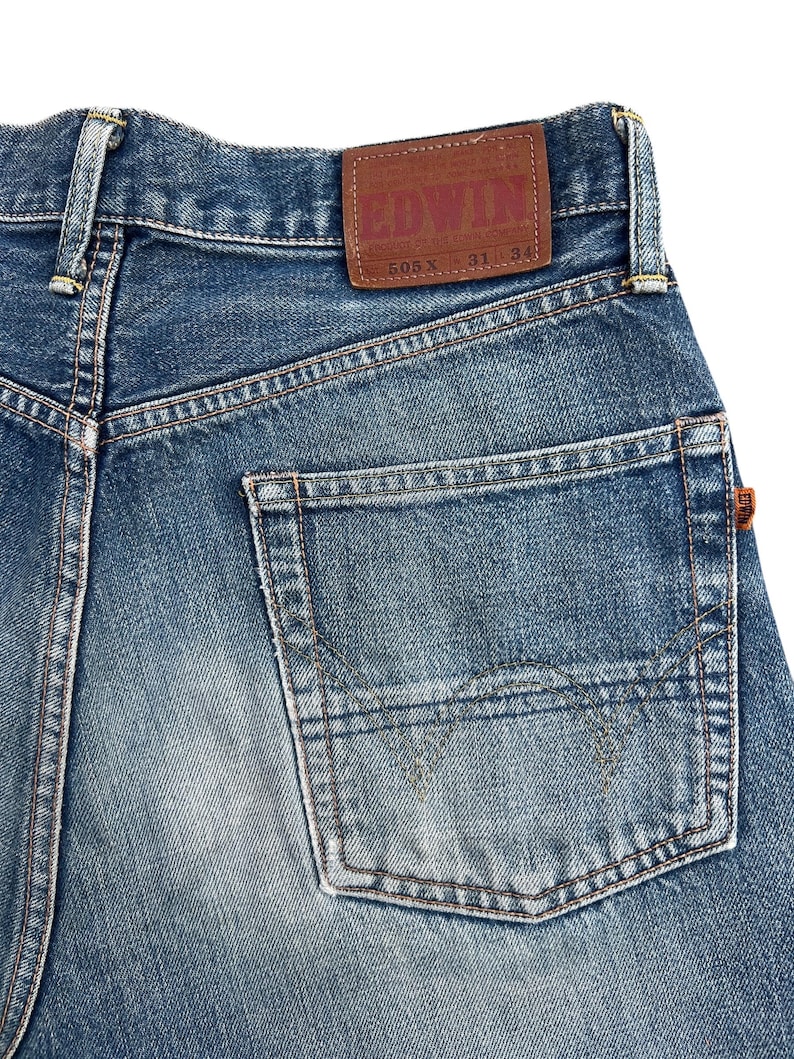 Puede incluir: Primer plano de la parte trasera de unos vaqueros de mezclilla azules. Los vaqueros presentan un parche de cuero marr&oacute;n con la marca "EDWIN" y detalles de talla. Se ve un bolsillo trasero, junto con costuras y la cl&aacute;sica textura de la mezclilla.