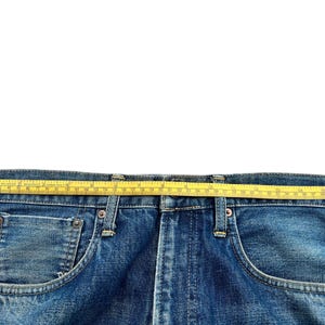 May include: Close-up of blue denim jeans with a yellow measuring tape across the waistband. The tape shows a measurement of approximately 16 inches. The jeans have classic five-pocket styling.