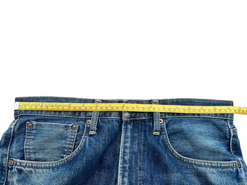 May include: Close-up of blue denim jeans with a yellow measuring tape across the waistband. The tape shows a measurement of approximately 16 inches. The jeans have classic five-pocket styling.
