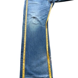 Pu&ograve; includere: Jeans in denim blu chiaro con lavaggio sbiadito e dettagli usurati. Un metro a nastro giallo &egrave; posizionato lungo la lunghezza dei jeans. I jeans hanno un classico design a cinque tasche e una chiusura con bottoni.