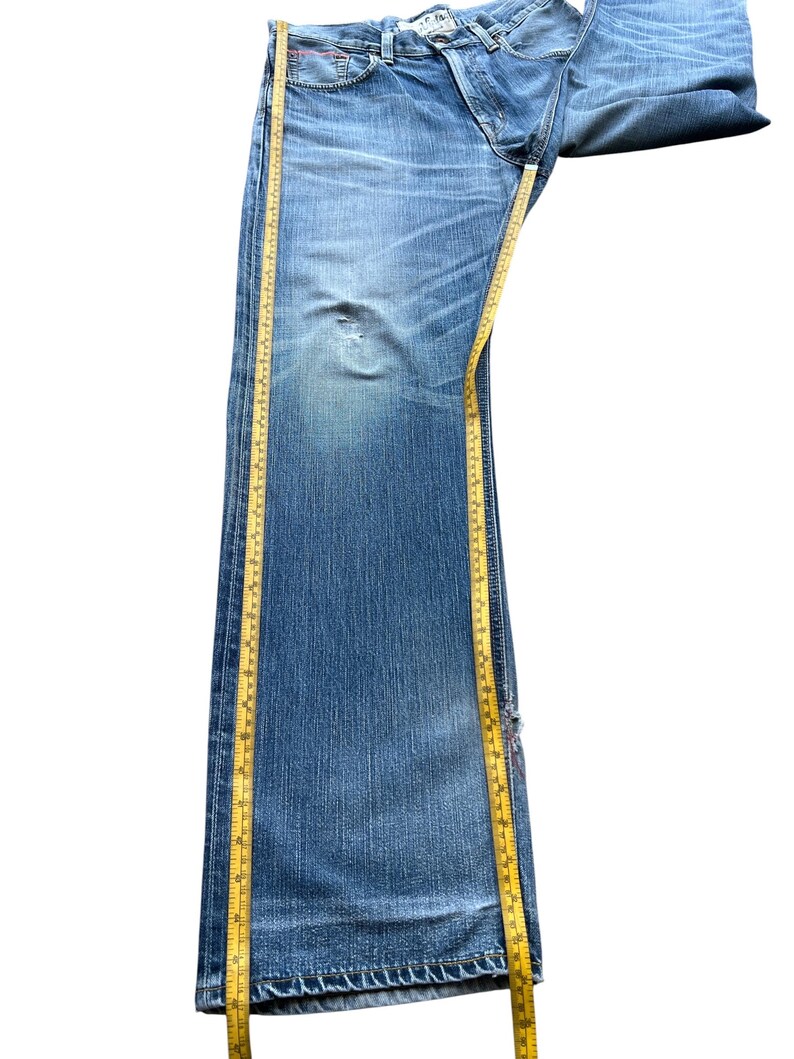 Pu&ograve; includere: Jeans in denim blu chiaro con lavaggio sbiadito e dettagli usurati. Un metro a nastro giallo &egrave; posizionato lungo la lunghezza dei jeans. I jeans hanno un classico design a cinque tasche e una chiusura con bottoni.