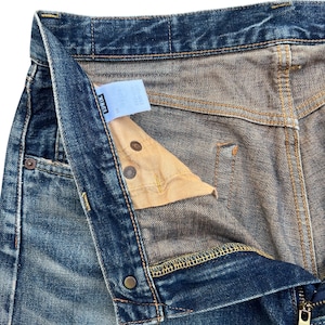 Puede incluir: Primer plano de unos vaqueros de mezclilla azules con la bragueta parcialmente desabrochada. Los vaqueros muestran signos de desgaste y decoloraci&oacute;n, con costuras visibles y botones met&aacute;licos. Una peque&ntilde;a etiqueta es visible dentro de la cintura.
