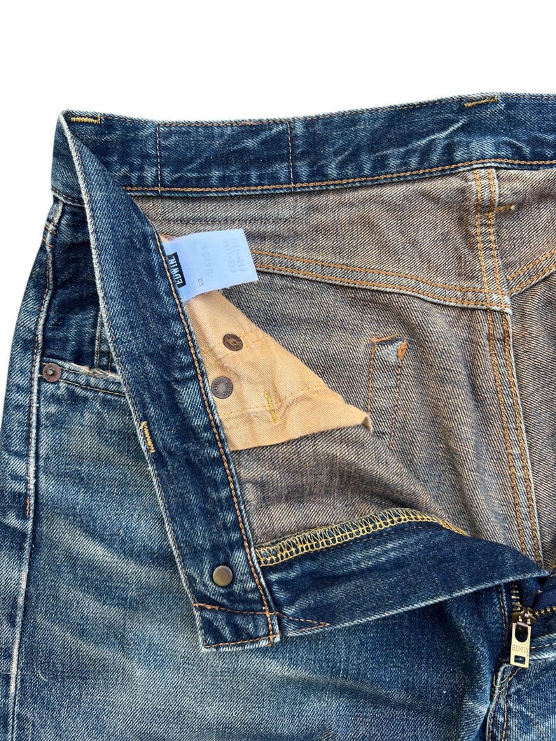 Puede incluir: Primer plano de unos vaqueros de mezclilla azules con la bragueta parcialmente desabrochada. Los vaqueros muestran signos de desgaste y decoloraci&oacute;n, con costuras visibles y botones met&aacute;licos. Una peque&ntilde;a etiqueta es visible dentro de la cintura.
