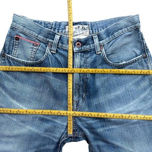Pu&ograve; includere: Jeans di denim blu chiaro con un lavaggio sbiadito. I jeans hanno un classico design a cinque tasche, una chiusura con bottone e cerniera e un piccolo accento rosso sulla tasca. Un metro a nastro giallo &egrave; posizionato sui jeans.