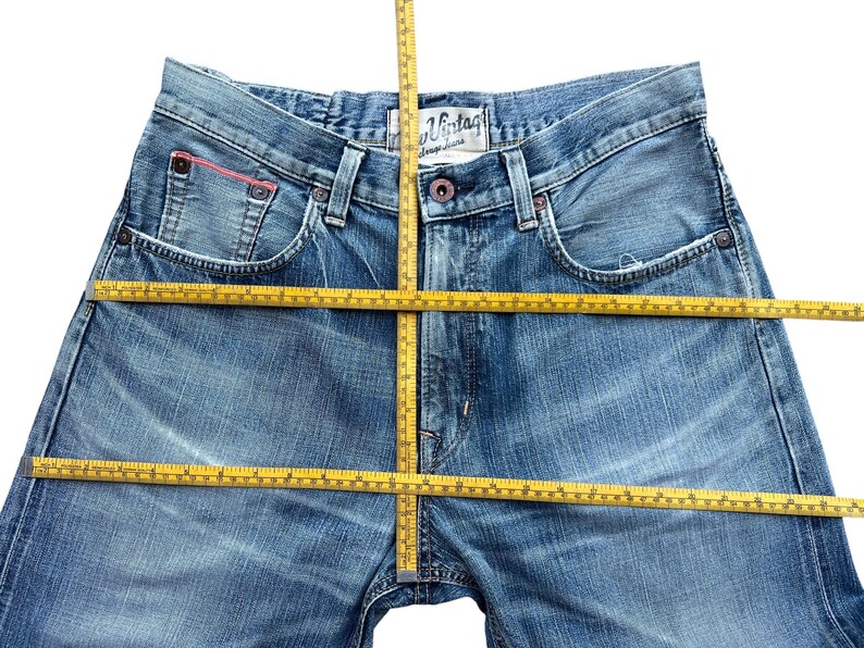 Pu&ograve; includere: Jeans di denim blu chiaro con un lavaggio sbiadito. I jeans hanno un classico design a cinque tasche, una chiusura con bottone e cerniera e un piccolo accento rosso sulla tasca. Un metro a nastro giallo &egrave; posizionato sui jeans.