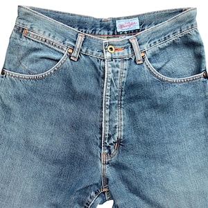 May include: Close-up of blue denim jeans with a classic five-pocket design. The jeans feature a button and zipper closure, belt loops, and the brand name "Wrangler" on a small tag. The denim has a faded wash, and the stitching is in a contrasting orange color.