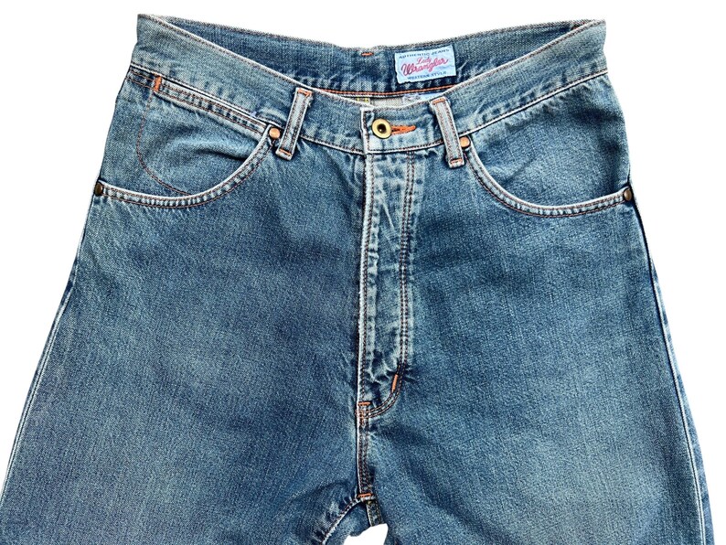 May include: Close-up of blue denim jeans with a classic five-pocket design. The jeans feature a button and zipper closure, belt loops, and the brand name "Wrangler" on a small tag. The denim has a faded wash, and the stitching is in a contrasting orange color.