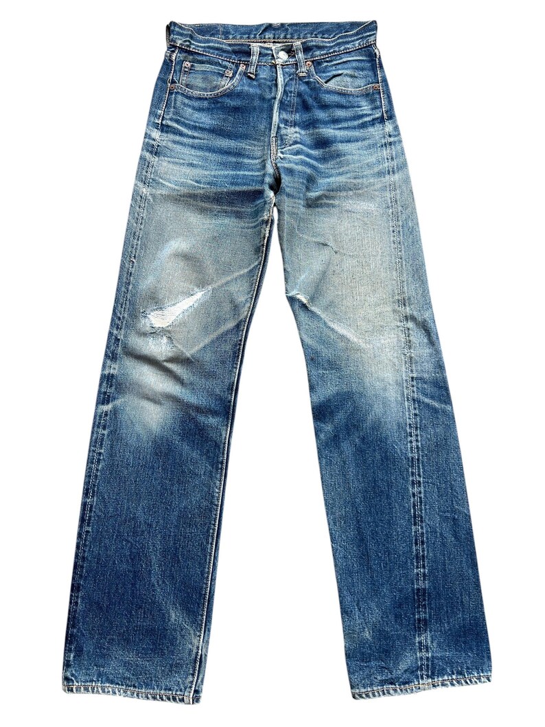Pu&ograve; includere: Jeans in denim blu sbiadito con chiusura a bottoni e classico design a cinque tasche. I jeans mostrano segni di usura con aree rovinate e un lavaggio chiaro, creando un'estetica vintage. I jeans sono a gamba dritta.