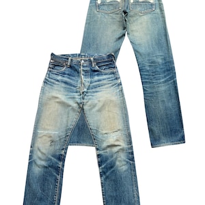May include: Two pairs of blue denim jeans. One pair is laid flat, showing the front with a button fly and distressed areas. The other pair is upright, displaying the back pockets with a white seagull design and a red leather patch.