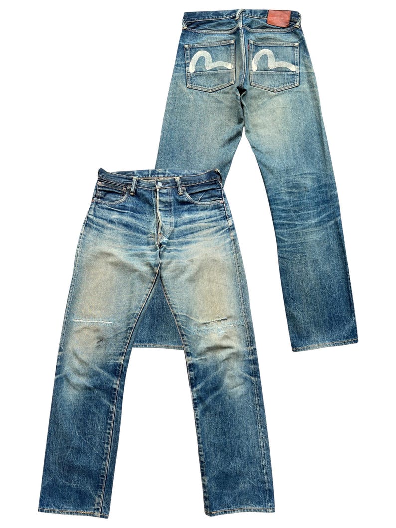 May include: Two pairs of blue denim jeans. One pair is laid flat, showing the front with a button fly and distressed areas. The other pair is upright, displaying the back pockets with a white seagull design and a red leather patch.