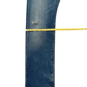 Puede incluir: Vaqueros azules de mezclilla con una zona de rodilla desgastada. Una cinta m&eacute;trica amarilla se coloca sobre los vaqueros para medir la longitud y el ancho. Los vaqueros est&aacute;n sobre un fondo blanco.