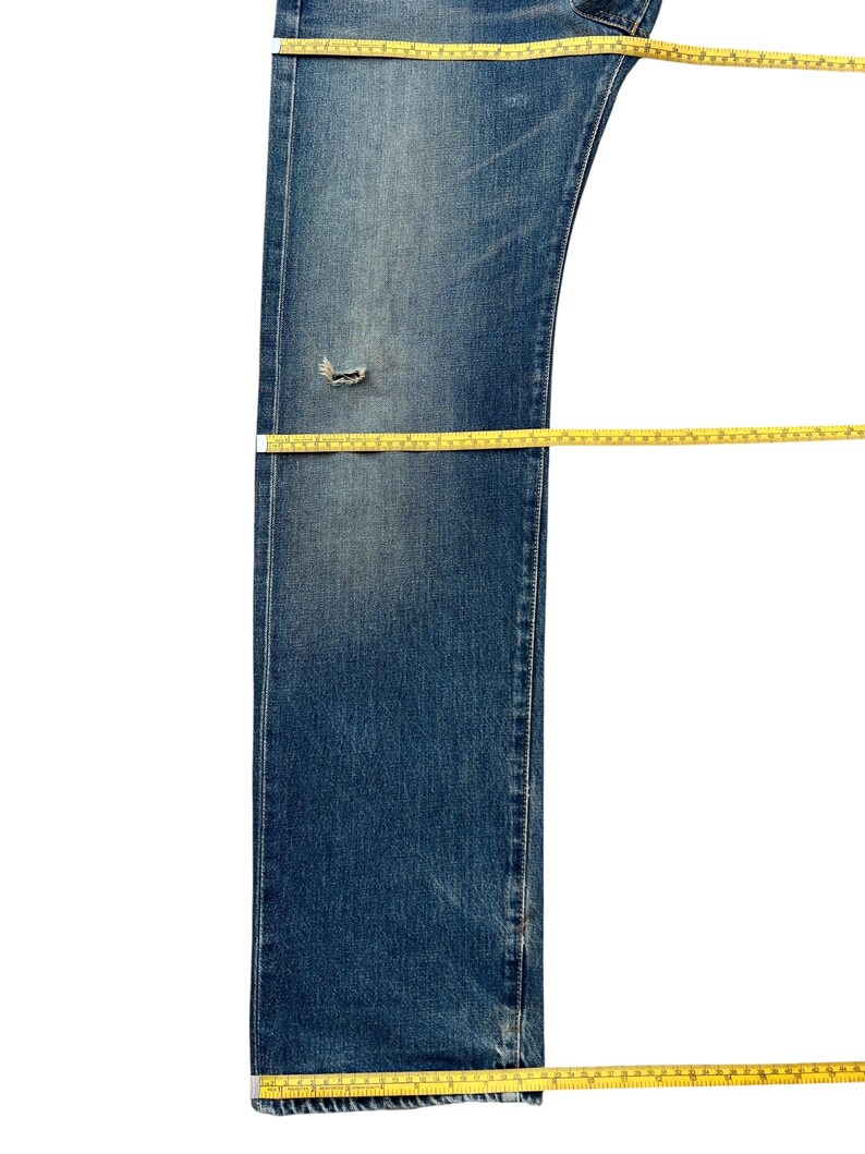 Puede incluir: Vaqueros azules de mezclilla con una zona de rodilla desgastada. Una cinta m&eacute;trica amarilla se coloca sobre los vaqueros para medir la longitud y el ancho. Los vaqueros est&aacute;n sobre un fondo blanco.