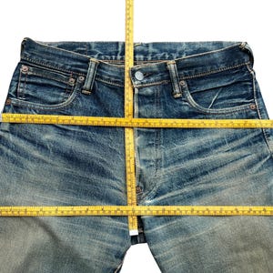 May include: Close-up of blue denim jeans with a yellow measuring tape laid across the waist and inseam. The jeans have a faded wash and visible wear marks, indicating a vintage or distressed style. The tape measures the garment's dimensions.