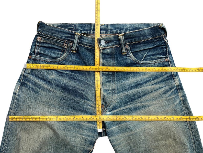 May include: Close-up of blue denim jeans with a yellow measuring tape laid across the waist and inseam. The jeans have a faded wash and visible wear marks, indicating a vintage or distressed style. The tape measures the garment's dimensions.