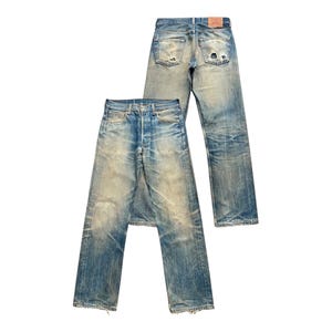 May include: Two pairs of vintage blue denim jeans with a faded wash and distressed details. One pair is laid flat, the other is upright. The jeans have a button fly and classic five-pocket design. The denim is worn and shows signs of wear.