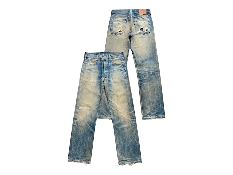 May include: Two pairs of vintage blue denim jeans with a faded wash and distressed details. One pair is laid flat, the other is upright. The jeans have a button fly and classic five-pocket design. The denim is worn and shows signs of wear.