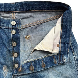 以下が含まれることがあります： ヴィンテージの青いデニムジーンズのクローズアップ。ボタンが外され、部分的に開いています。ジーンズは、色あせた部分やダメージのあるパッチなど、使用感が見られます。シルバーの金属ボタンとリベット、ベージュの内側の裏地が見えます。