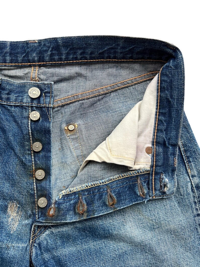 以下が含まれることがあります： ヴィンテージの青いデニムジーンズのクローズアップ。ボタンが外され、部分的に開いています。ジーンズは、色あせた部分やダメージのあるパッチなど、使用感が見られます。シルバーの金属ボタンとリベット、ベージュの内側の裏地が見えます。