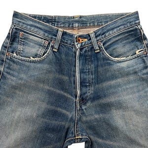 Puede incluir: Primer plano de unos vaqueros de mezclilla azules. Los vaqueros tienen bragueta con botones, dise&ntilde;o cl&aacute;sico de cinco bolsillos y muestran signos de desgaste con zonas descoloridas y detalles desgastados. Las costuras son visibles y el aspecto general es vintage.