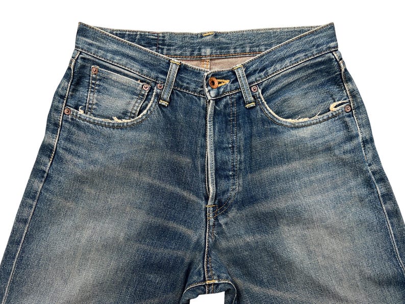 Puede incluir: Primer plano de unos vaqueros de mezclilla azules. Los vaqueros tienen bragueta con botones, dise&ntilde;o cl&aacute;sico de cinco bolsillos y muestran signos de desgaste con zonas descoloridas y detalles desgastados. Las costuras son visibles y el aspecto general es vintage.