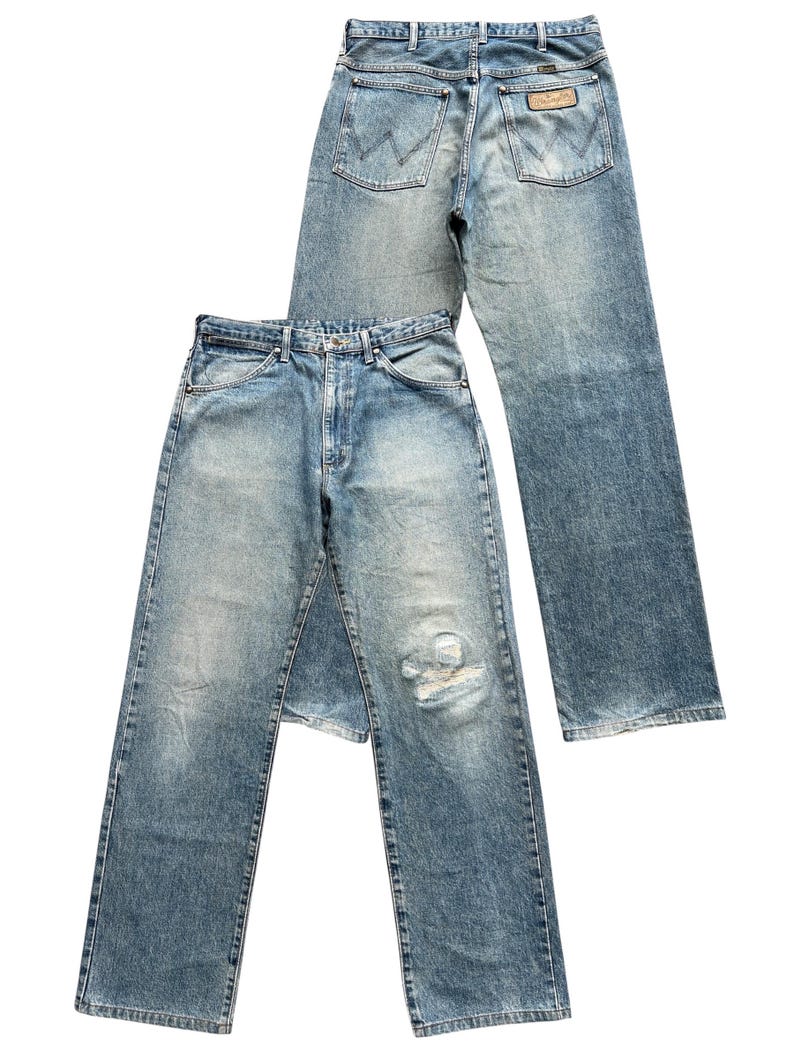 K&ouml;nnte beinhalten: Zwei Paar Vintage-Jeans aus blauem Denim. Ein Paar liegt &uuml;ber dem anderen. Die Jeans haben ein klassisches F&uuml;nf-Taschen-Design, eine verblasste Waschung und einen abgenutzten Bereich an der Vorderseite eines Beins. Die Ges&auml;&szlig;taschen weisen ein markantes Nahtmuster auf.