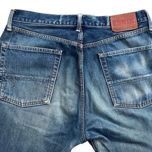 Puede incluir: Primer plano de la parte trasera de unos vaqueros azules. Los vaqueros tienen dos bolsillos traseros y un parche de cuero marr&oacute;n con la marca "EDWIN". El denim tiene un aspecto descolorido y desgastado.