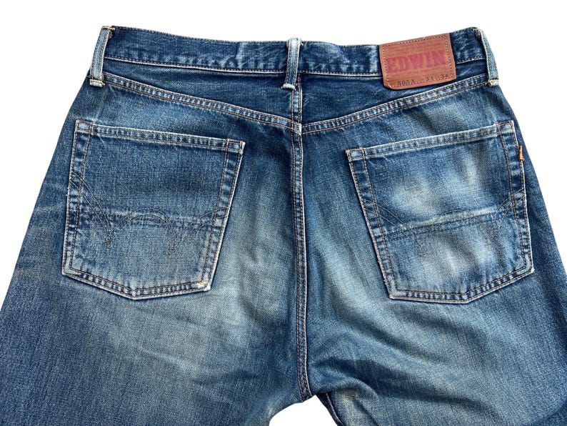 Puede incluir: Primer plano de la parte trasera de unos vaqueros azules. Los vaqueros tienen dos bolsillos traseros y un parche de cuero marr&oacute;n con la marca "EDWIN". El denim tiene un aspecto descolorido y desgastado.