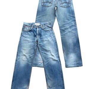 Pu&ograve; includere: Due paia di jeans in denim blu chiaro. Un paio &egrave; steso, mostrando la parte anteriore con la chiusura a bottoni e le tasche. L'altro paio &egrave; in piedi, mostrando le tasche posteriori e una toppa in pelle. Entrambi i jeans hanno un aspetto usurato e sbiadito.