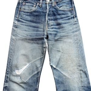 Pu&ograve; includere: Jeans di denim blu sbiadito con chiusura a bottoni e classico design a cinque tasche. I jeans presentano dettagli usurati, tra cui uno strappo sul ginocchio sinistro. Il denim mostra segni di usura e sbiadimento.
