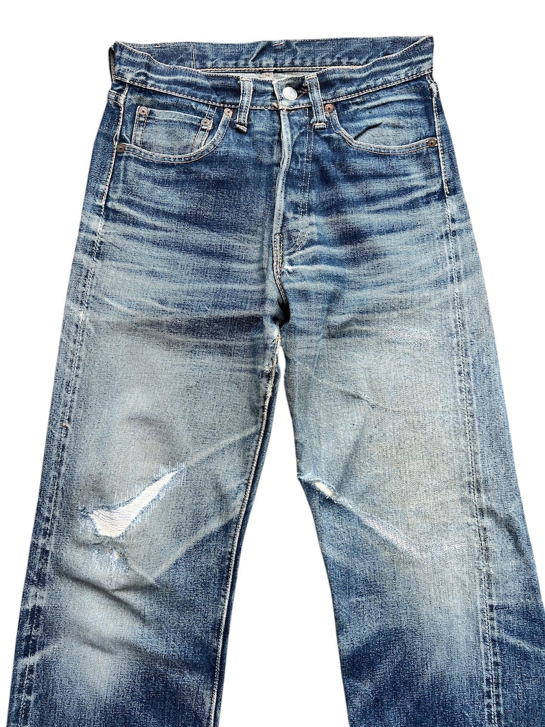 Pu&ograve; includere: Jeans di denim blu sbiadito con chiusura a bottoni e classico design a cinque tasche. I jeans presentano dettagli usurati, tra cui uno strappo sul ginocchio sinistro. Il denim mostra segni di usura e sbiadimento.