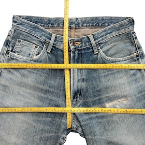 May include: Faded blue denim jeans with a classic five-pocket design. A yellow measuring tape is laid across the waist and down the inseam, showing the dimensions of the jeans. The jeans have a worn, distressed look.