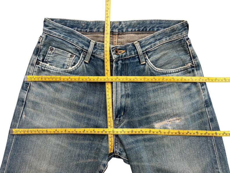 May include: Faded blue denim jeans with a classic five-pocket design. A yellow measuring tape is laid across the waist and down the inseam, showing the dimensions of the jeans. The jeans have a worn, distressed look.