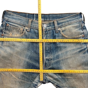 May include: Close-up of blue denim jeans with a button closure and pockets. A yellow measuring tape is laid across the jeans to measure the waist and inseam. The jeans have a worn, faded appearance.