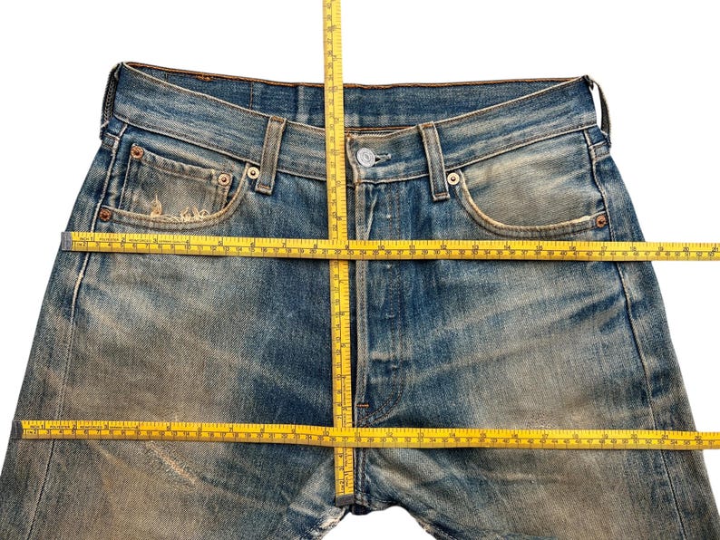 May include: Close-up of blue denim jeans with a button closure and pockets. A yellow measuring tape is laid across the jeans to measure the waist and inseam. The jeans have a worn, faded appearance.