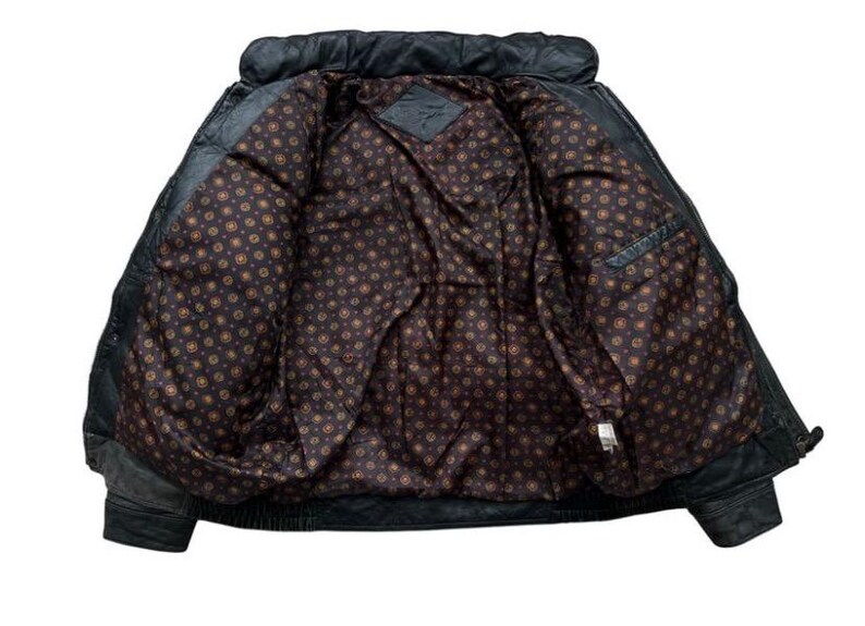 May include: Black leather jacket with a quilted collar and a patterned interior lining. The jacket has a classic design with a front zipper and pockets. The interior lining features a repeating pattern of small circles in shades of brown and gold.