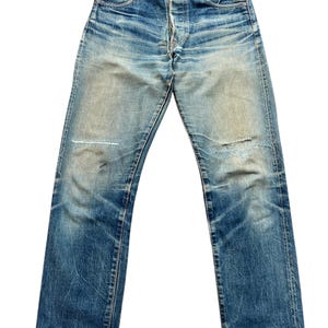 May include: Faded blue denim jeans with a button fly and classic five-pocket design. The jeans show signs of wear, including a patched knee and distressed areas, giving them a vintage look. The jeans are a straight-leg style.