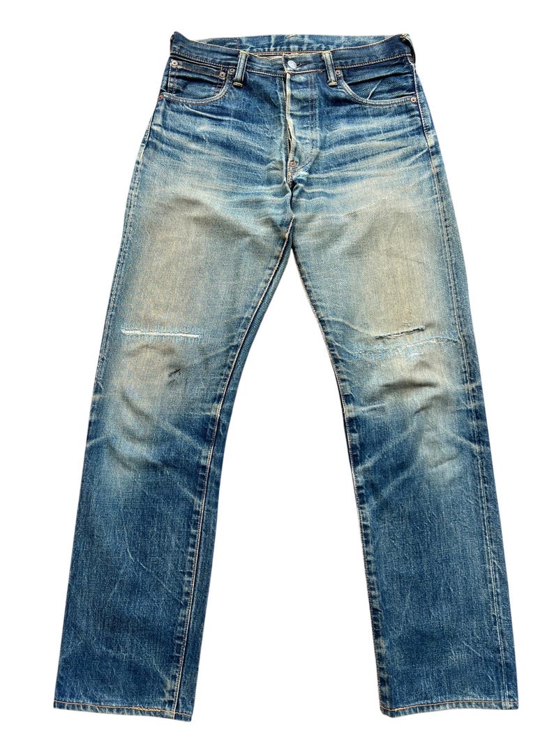 May include: Faded blue denim jeans with a button fly and classic five-pocket design. The jeans show signs of wear, including a patched knee and distressed areas, giving them a vintage look. The jeans are a straight-leg style.