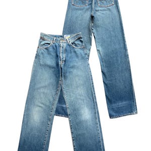 May include: Two pairs of light blue denim jeans. One pair is laid flat, showing the front with a button closure and pockets. The other pair is behind it, showing the back with pockets and a brown leather patch.