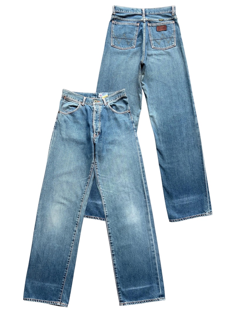 May include: Two pairs of light blue denim jeans. One pair is laid flat, showing the front with a button closure and pockets. The other pair is behind it, showing the back with pockets and a brown leather patch.