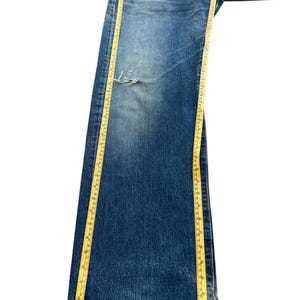 Puede incluir: Vaqueros de mezclilla azul descolorido con un aspecto desgastado, con un desgarro en la parte delantera. Una cinta m&eacute;trica amarilla se coloca a lo largo del pantal&oacute;n. Los vaqueros parecen ser vintage o pre-usados.