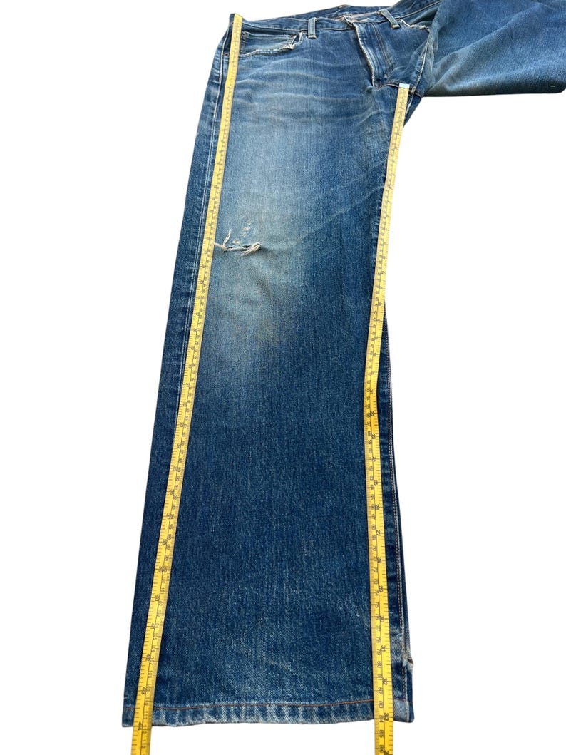 Puede incluir: Vaqueros de mezclilla azul descolorido con un aspecto desgastado, con un desgarro en la parte delantera. Una cinta m&eacute;trica amarilla se coloca a lo largo del pantal&oacute;n. Los vaqueros parecen ser vintage o pre-usados.