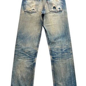 May include: Faded blue denim jeans with distressed details, including rips and wear marks. The back pockets and a red tag are visible. The jeans have a classic five-pocket design and a straight leg cut.