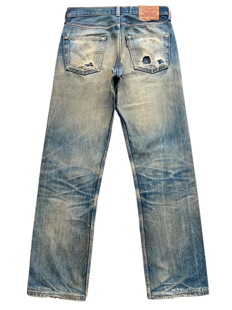 May include: Faded blue denim jeans with distressed details, including rips and wear marks. The back pockets and a red tag are visible. The jeans have a classic five-pocket design and a straight leg cut.