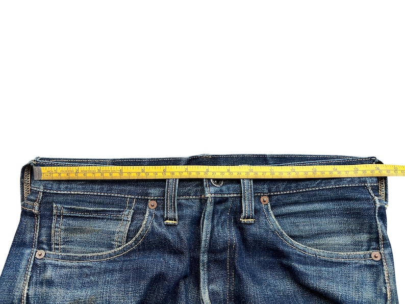 Może przedstawiać: Zbliżenie na pas niebieskich dżins&oacute;w z ż&oacute;łtą miarką ułożoną na g&oacute;rze. Dżinsy mają zapięcie na guziki, kieszenie i szlufki. Miarka pokazuje pomiar w talii w centymetrach.