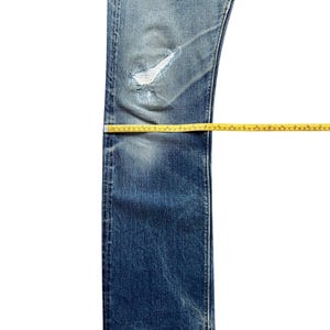 Pu&ograve; includere: Jeans di denim blu dall'aspetto usurato, con un ginocchio strappato. Un metro a nastro giallo viene utilizzato per misurare la lunghezza e la larghezza dei jeans. I jeans sono su sfondo bianco.