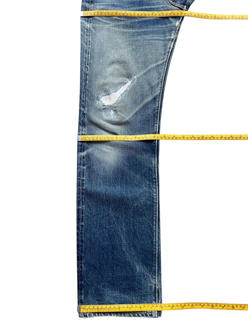 Pu&ograve; includere: Jeans di denim blu dall'aspetto usurato, con un ginocchio strappato. Un metro a nastro giallo viene utilizzato per misurare la lunghezza e la larghezza dei jeans. I jeans sono su sfondo bianco.