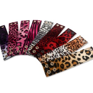May include: A collection of seven animal print wrist cuffs in various colors, including purple, pink, red, and brown. Each cuff features a snap closure with gold-colored metal hardware. The cuffs are arranged in a fan shape against a white background.