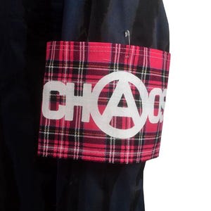Pode incluir: Uma braçadeira xadrez rosa e preta com a palavra "CHAOS" em letras brancas e um símbolo de anarquia branco. A braçadeira é presa a uma peça de roupa preta com um alfinete de segurança.