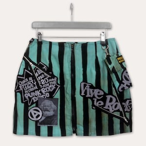 Op de afbeelding: Een turquoise en zwart gestreepte minirok met een ritssluiting aan de voorkant en meerdere patches. Patches bevatten de tekst "OURS IS EFFORT LEAVE IN ORDER PUNK ROCK", "Vive le Rock" en een portret. De rok heeft een klein aangehecht zakje.